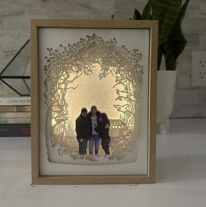 A custom family portrait lightbox. The centerpiece showcases a beautiful family portrait, while layers of carefully crafted cardstock create a lush and vibrant woodland setting. Ideal for unique home decor & personalized gifts.